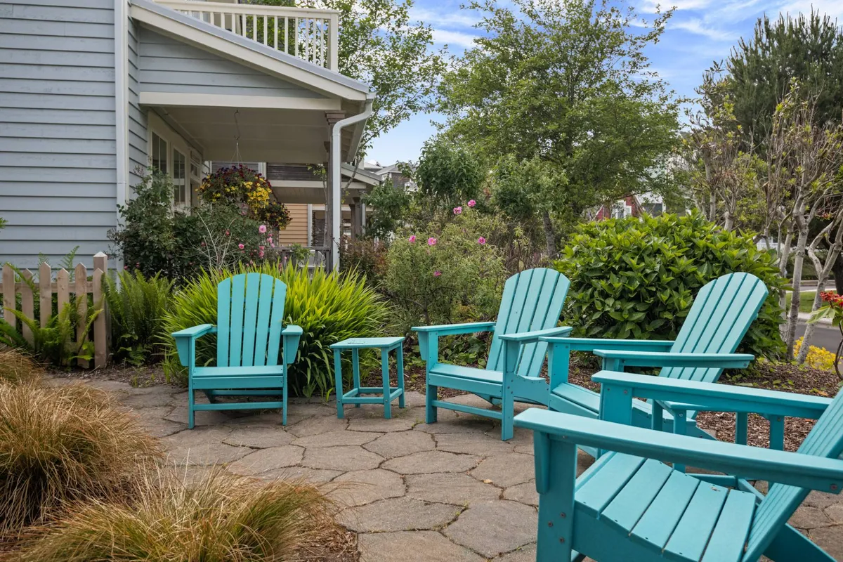 A cheerful stone patio scene with turquoise Adirondack chairs and lush greenery, bathed in morning light. The composition emphasizes peaceful outdoor moments and a connection to the community's natural surroundings.