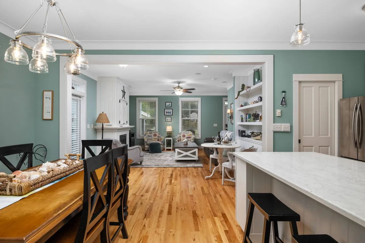 A wide, inviting view of the open-concept living, dining, and kitchen area. The dining table is the focal point, surrounded by a comfortable sofa in the living area and a kitchen island nearby. Bookshelves and curated décor add coastal charm, while sunlight illuminates the entire space, creating a warm and welcoming atmosphere.