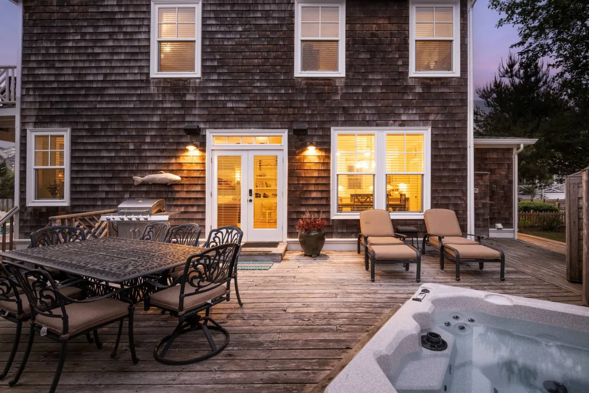 This exterior image captures the back of the house, featuring a welcoming deck with a large dining table, a bubbling hot tub, a grill, and the back door leading inside. The scene is set for outdoor meals, hot tub relaxation, and casual gatherings, with soft sunset light enhancing the friendly, laid-back mood.