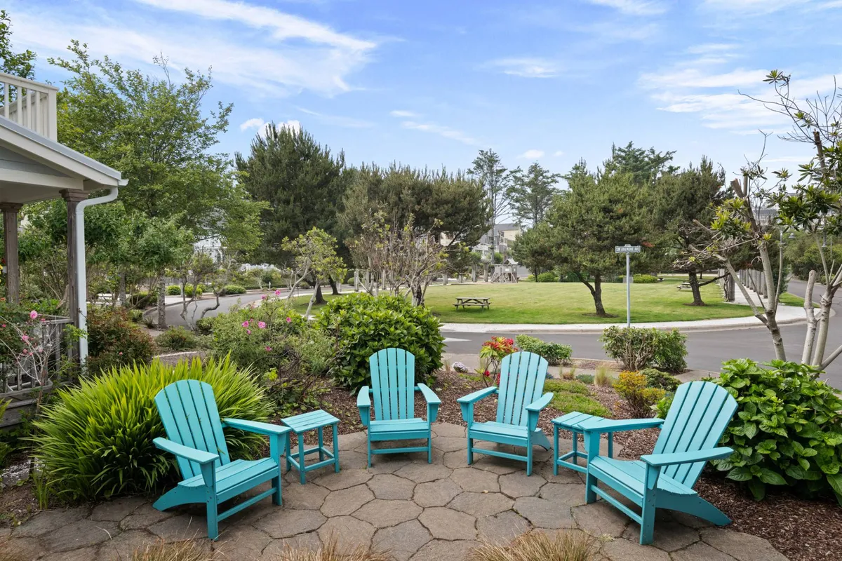 This exterior image features turquoise Adirondack chairs arranged around a stone patio, surrounded by lush green landscaping. The scene invites relaxation and socializing just outside the home, with a clear sense of the community’s outdoor charm.