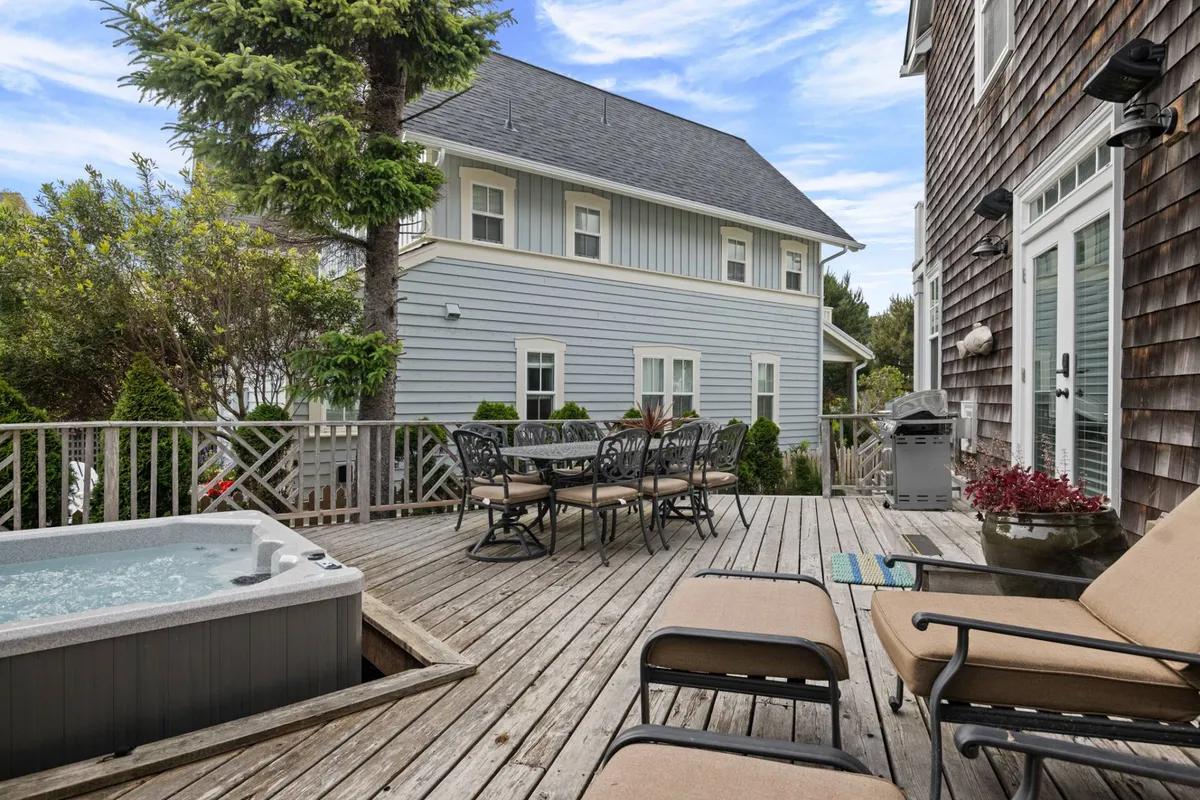 A vibrant exterior shot of the wooden deck surrounded by lush greenery, with a hot tub and outdoor dining set as focal points. The setup encourages both relaxation in the hot tub and group meals or evening chats at the table, all framed by a tranquil natural setting.
