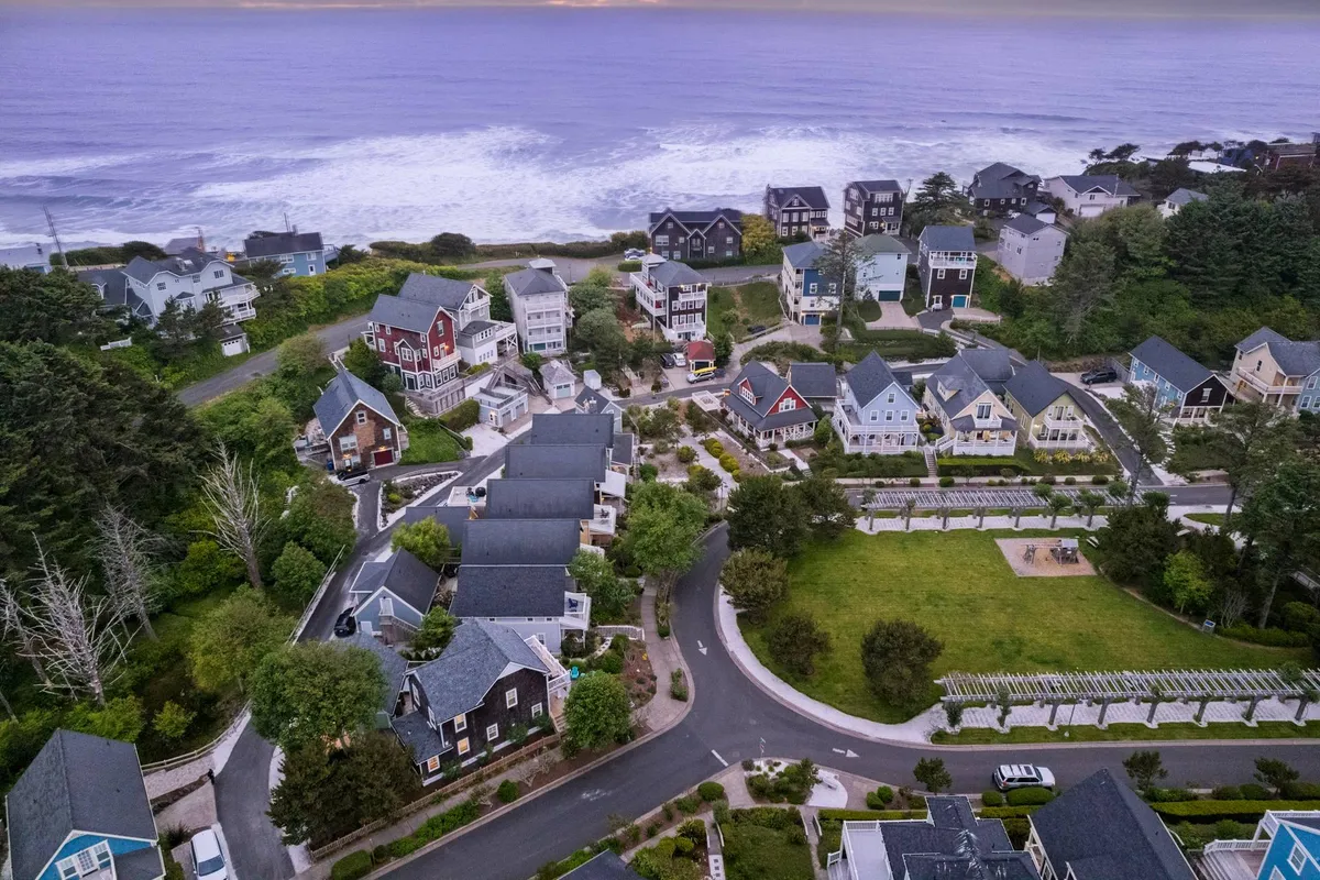The image captures a serene coastal view with gentle ocean waves, colorful beach houses, and a vibrant green park area directly across the street. The park features open lawns and play spaces, inviting families for gatherings, picnics, and outdoor fun while the ocean sparkles in the background.