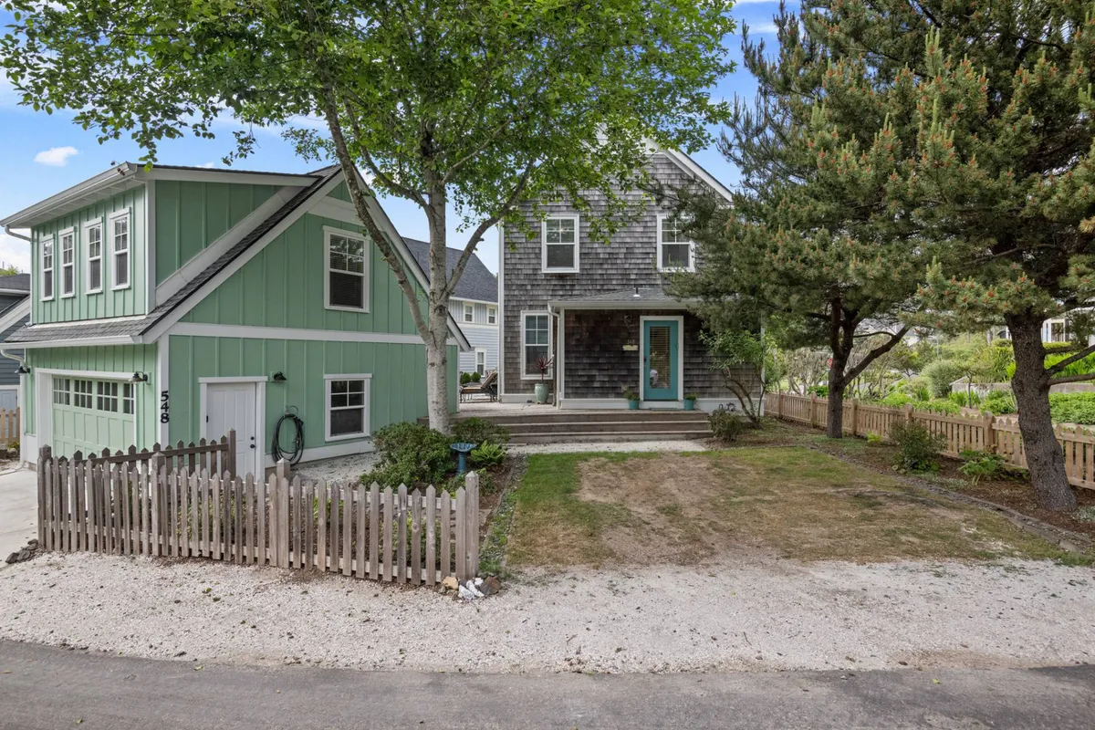 This image features a welcoming two-story house with a green garage, spacious front porch, and a large fenced yard shaded by mature trees—ideal for family gatherings and outdoor play. The scene evokes morning coffees on the porch, relaxed evenings, and the possibility of kids playing safely within the yard.