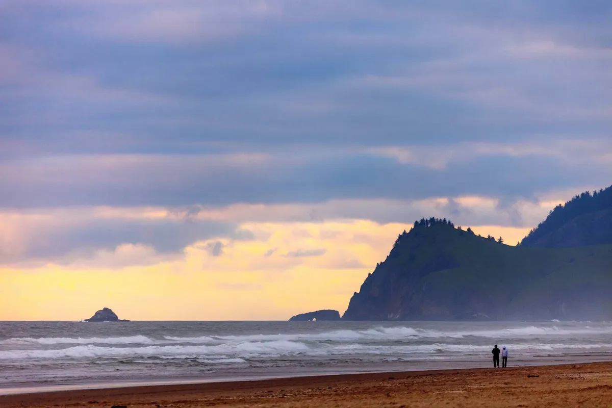 A dramatic oceanfront scene with rolling waves, distant cliffs, and a couple strolling along the shoreline. The mood is romantic and tranquil, and the private beachfront access is visually emphasized, highlighting the unique perk of direct connection to the coast.