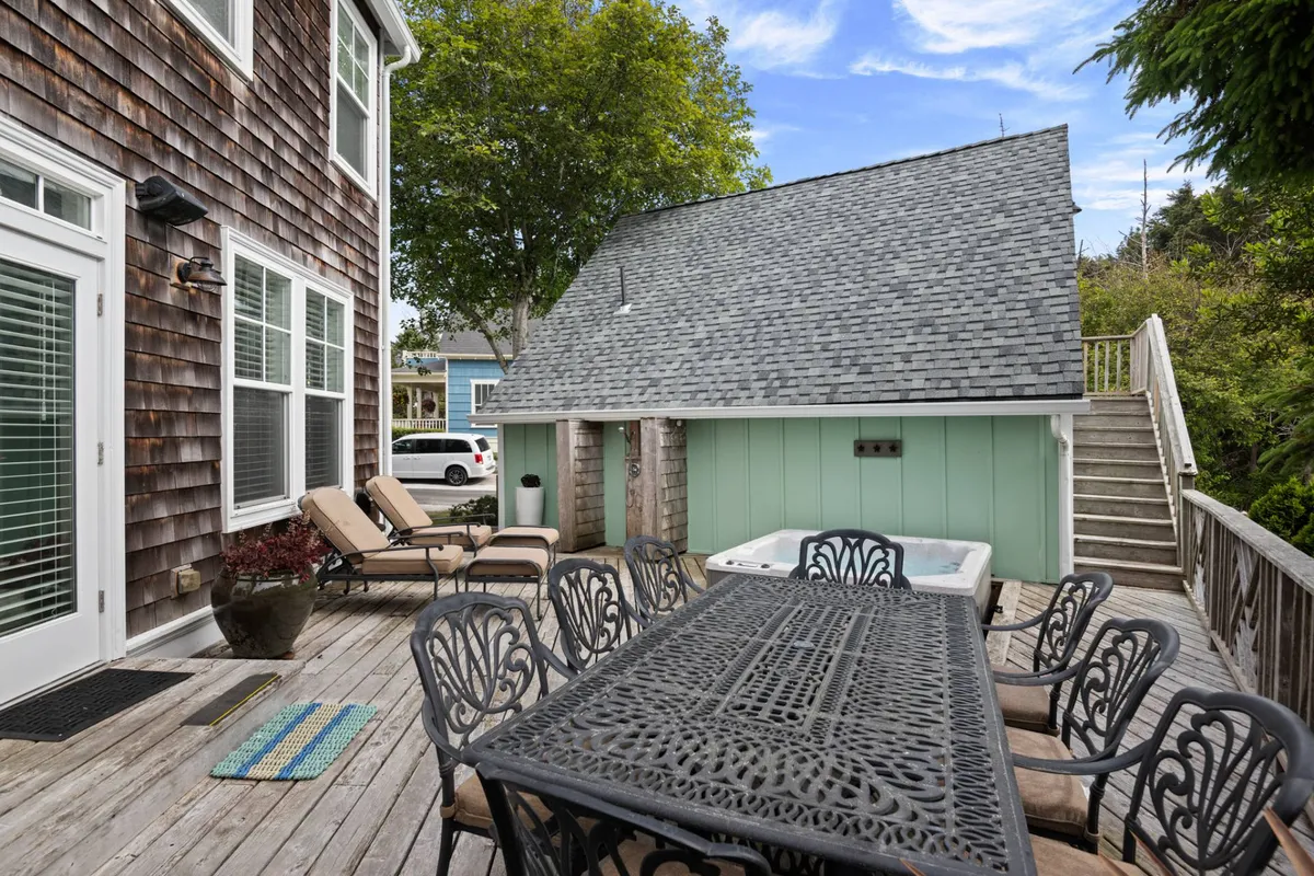 Captured from a different angle, this image highlights a lively outdoor space with a wooden deck, dining table, hot tub, lounge chairs, and lush greenery. The area is set up for gatherings, sunbathing, and enjoying meals in the open air, with trees providing natural shade and privacy.