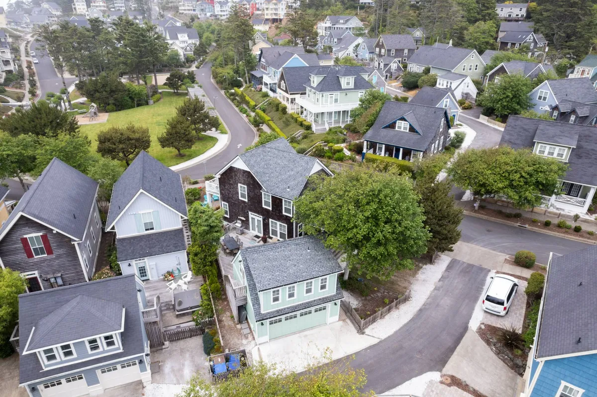 A sweeping view of the Olivia Beach neighborhood, showcasing diverse architectural styles of the homes, lush green trees, and curved residential roads. The scene is lively yet peaceful, with ample green space, signs of community interaction, and children playing, giving a sense of the storybook setting described.
