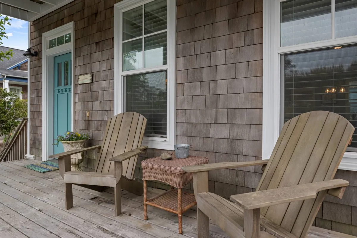 A cozy front porch scene with two wooden Adirondack chairs and a small table, set against a backdrop of a blue front door and classic wooden shingle siding. The image captures an inviting spot for quiet moments—perfect for sipping morning coffee or relaxing with a partner or close friends.