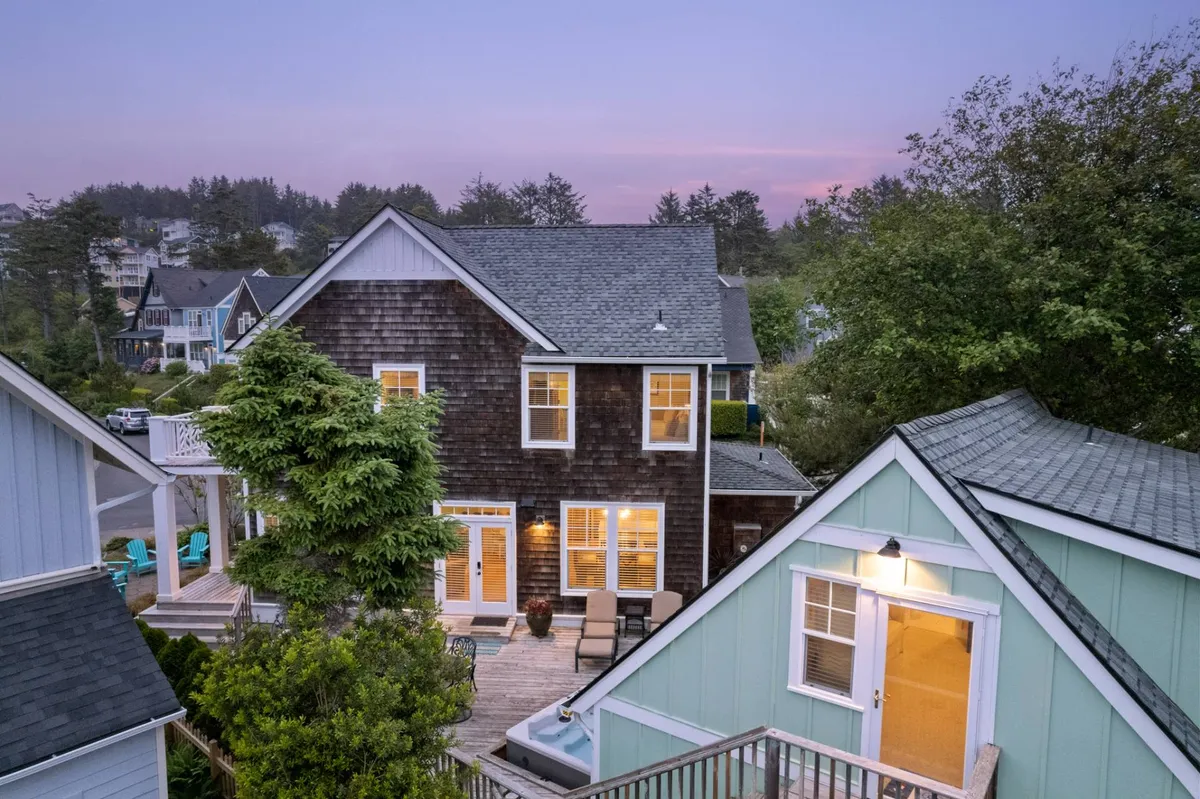 The exterior showcases a charming two-story home with warm wooden siding, a leafy green tree in the foreground, and a deck area equipped with chairs. This image radiates peacefulness and an inviting atmosphere, highlighting both the home and its connection to lush, natural surroundings.