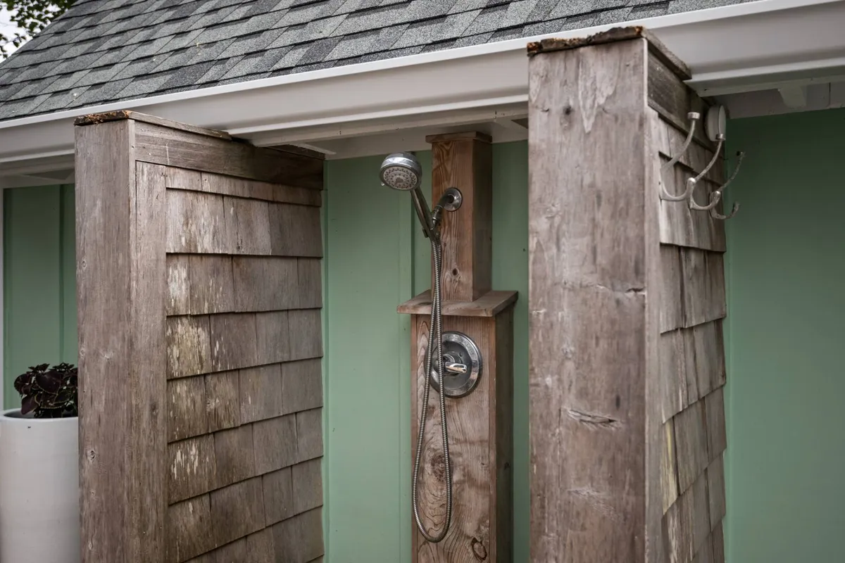 This serene image shows an outdoor shower with weathered wood panels and a green wall, surrounded by natural elements that evoke a peaceful connection to the outdoors. The shower space feels inviting yet private—perfect for rinsing off after a beach day or hot tub soak.