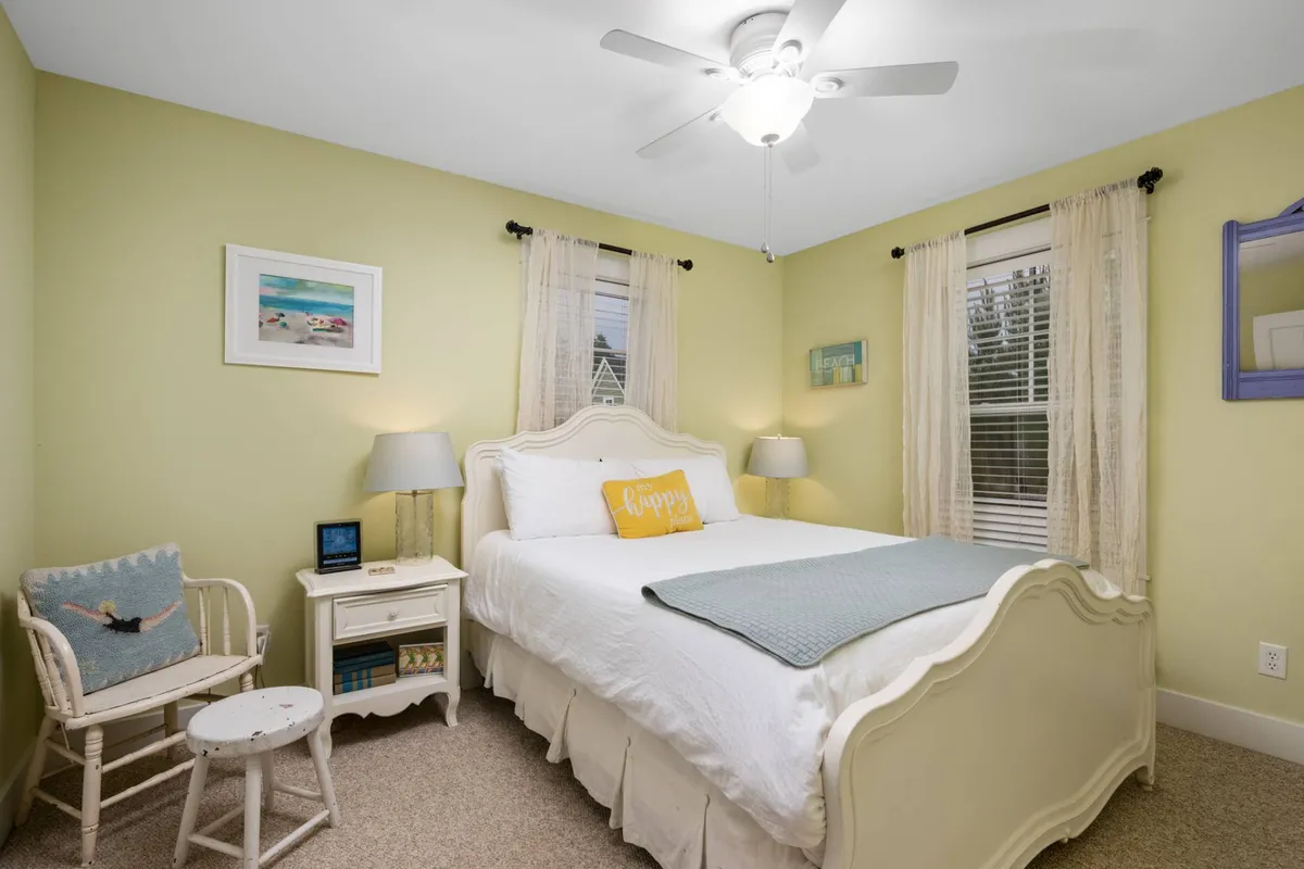 A calm and happy bedroom scene featuring a bed with decorative pillows, a nightstand with a lamp, and framed artwork on the wall. Natural light streams in, showcasing a tranquil and cheerful environment.
